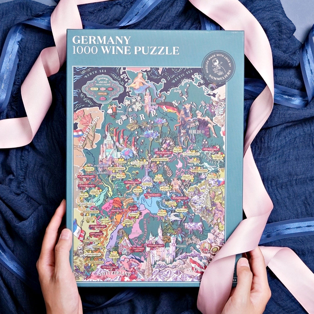 Geschlossene Puzzle-Box "Germany 1000 Wine Puzzle", gehalten von Händen, umrahmt von einem dunkelblauen Tuch und einem rosa Satinband.