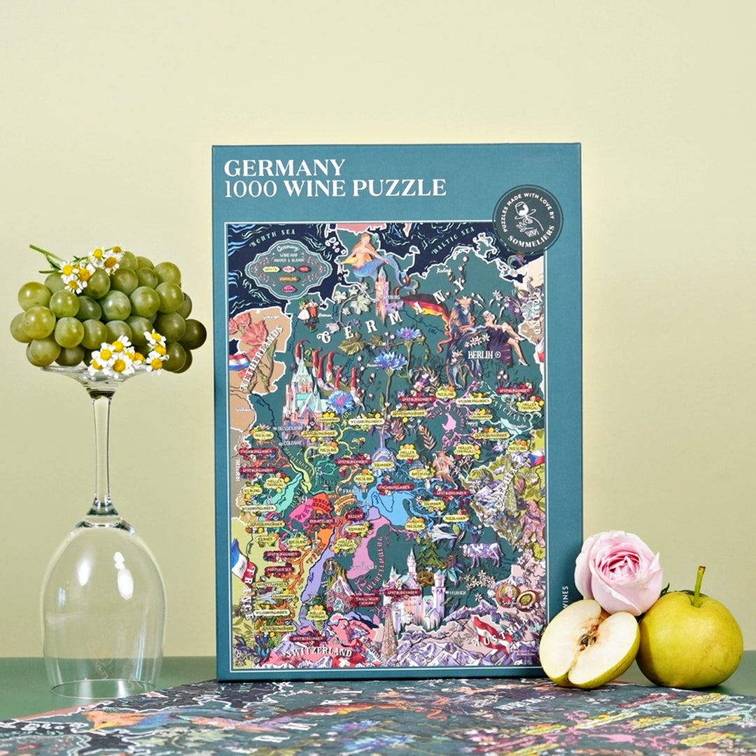 Produktaufnahme der Puzzle-Box "Germany 1000 Wine Puzzle" neben einem umgedrehten Weinglas, das mit Trauben und Gänseblümchen dekoriert ist, sowie einer halbierten Birne und einer Rose.\