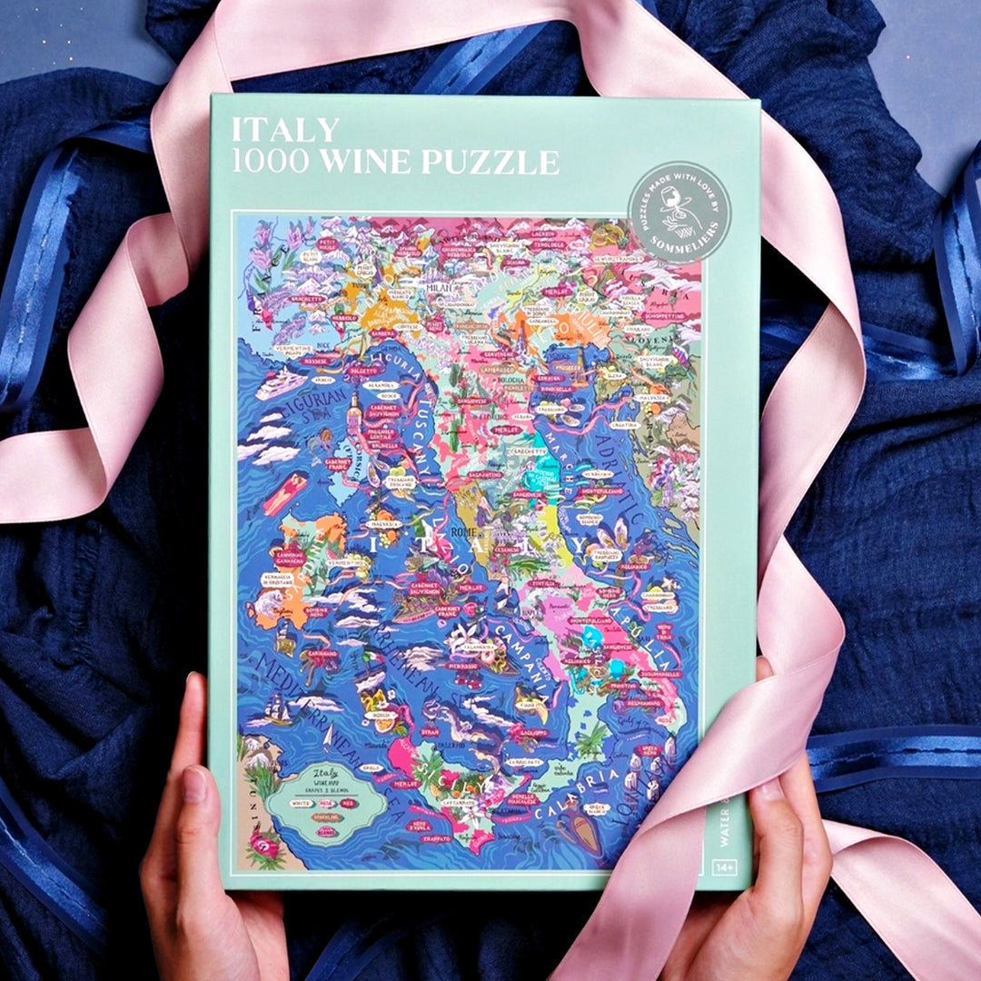 Geschlossene Puzzle-Box "Italy 1000 Wine Puzzle", gehalten von Händen, umrahmt von einem dunkelblauen Tuch und einem rosa Satinband.