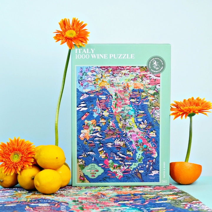 Produktaufnahme der Italien-Puzzle-Box, umgeben von gelben Zitronen, einer halben Orange und orangen Gerbera-Blumen.