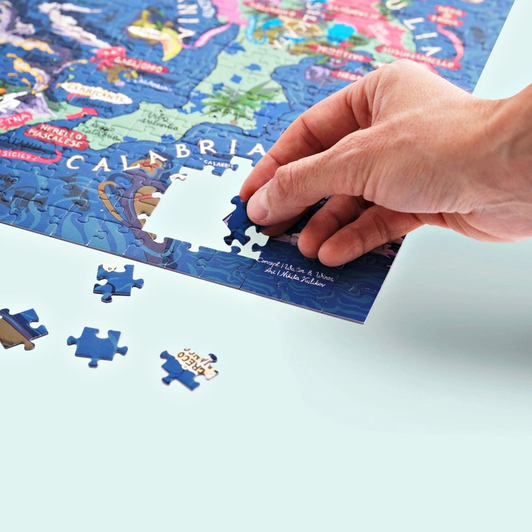 Nahaufnahme einer Hand, die ein Puzzleteil in die Region Kalabrien einfügt; der Name "Greco" ist auf einem losen Puzzleteil sichtbar.