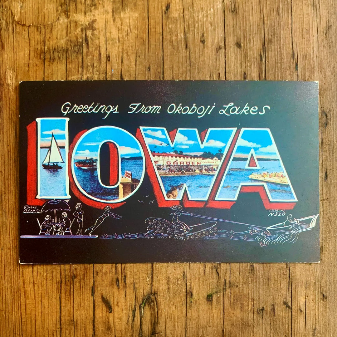 Dunkle Vintage-Postkarte "Greetings From Okoboji Lakes IOWA". Die Buchstaben zeigen Seemotive und das Ufer, mit stilisierten Zeichnungen von Menschen, die schwimmen und sich sonnen, im Vordergrund.