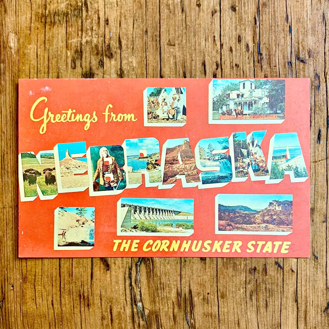 Vintage-Postkarte "Greetings from NEBRASKA THE CORNHUSKER STATE" auf leuchtend rotem Hintergrund. Der Schriftzug ist mit Motiven von Büffeln, historischen Figuren und Dämmen gefüllt.